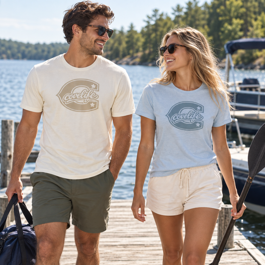 A man and a woman walking on a wooden dock by the water, each wearing a short-sleeve graphic T-shirt. The man's shirt is cream with a muted taupe logo, and the woman's shirt is light blue with a muted blue logo.