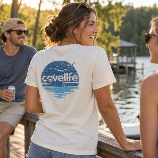 A woman standing on a wooden dock by a lake wears a cream-colored t-shirt with a blue circular graphic on the back that reads 'covelife'. A man in a blue shirt and sunglasses sits to her left, and another person is partially visible on the right, with trees and a dock in the background.