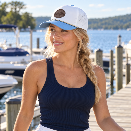 A woman wearing a white and light blue trucker-style cap with a covelife leather patch on the front, paired with a navy ribbed tank top, standing on a wooden dock with a lake and boats in the background.