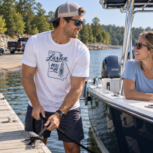 A man wearing a white graphic t-shirt with a 'Lamer Deal Day' print and a gray cap is docking a boat on a wooden pier next to a woman on a white motorboat.