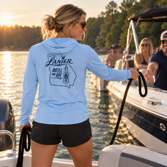 A woman on a boat at sunset wearing a light blue long-sleeve hooded shirt with a dark graphic print on the back that reads 'LAKE LANIER' and 'DEAL ME IN'.