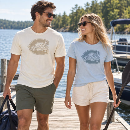 A man and a woman walking on a wooden dock by the water, each wearing a short-sleeve graphic T-shirt. The man's shirt is cream with a muted taupe logo, and the woman's shirt is light blue with a muted blue logo.