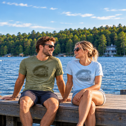 A man and a woman sitting on a wooden dock by a lake, both wearing matching graphic t-shirts with the brand name 'COVELiFE' printed on the chest. The man wears an olive green shirt and the woman wears a light blue shirt, with a forested shoreline and blue sky in the background.