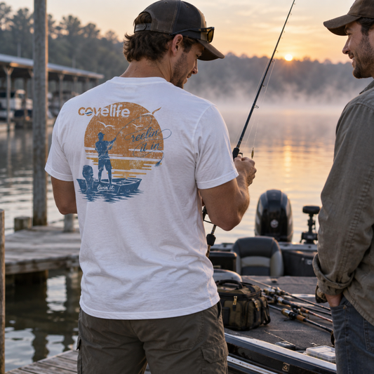 A man standing on a boat at a lake dock at sunrise, wearing a white crewneck t-shirt with a large back graphic that reads 'covelife' "reelin it in" and 'recreation on the water'.