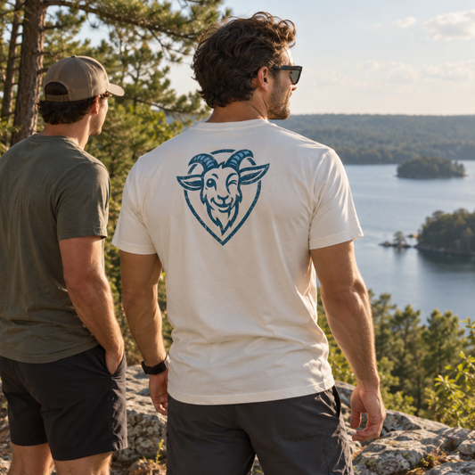 Two men standing outdoors on a rocky overlook with a lake and forest in the background. The man in the center is shown from the back wearing a white T-shirt with a blue ram/goat head graphic on the upper back.