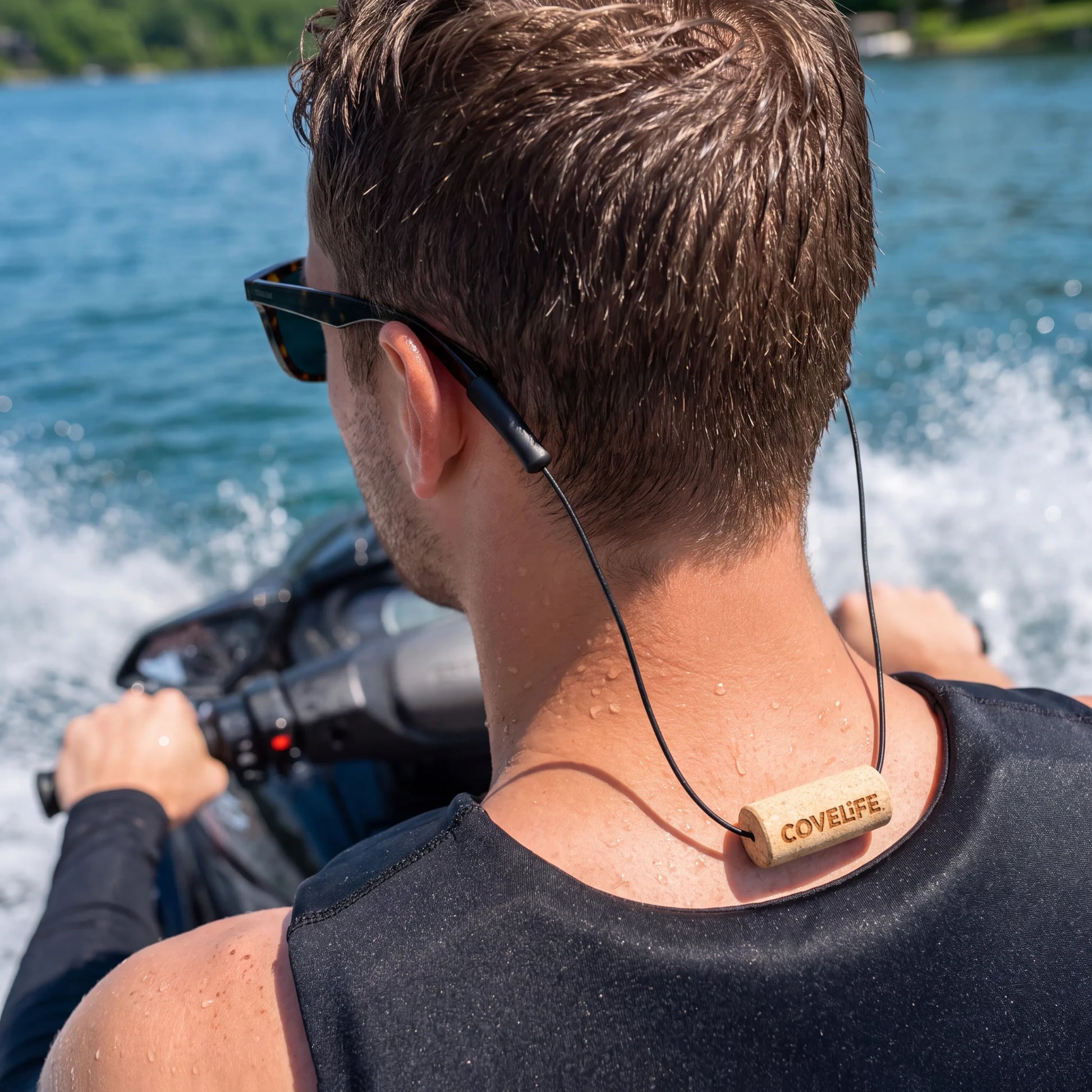 A man on a jet ski seen from behind wearing a black wetsuit top and sunglasses, with the COVELiFE sunglass retainer against a blue water background.
