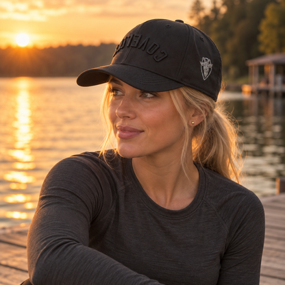 A woman wearing a black baseball cap with COVELiFE tonal embroidery photographed outdoors by a lake at golden hour sunset. 