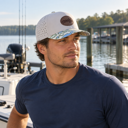 A man wearing a white and blue patterned baseball cap with a brown oval patch that reads COVELiFE on the brim, photographed outdoors at a dock with water and trees in the background.