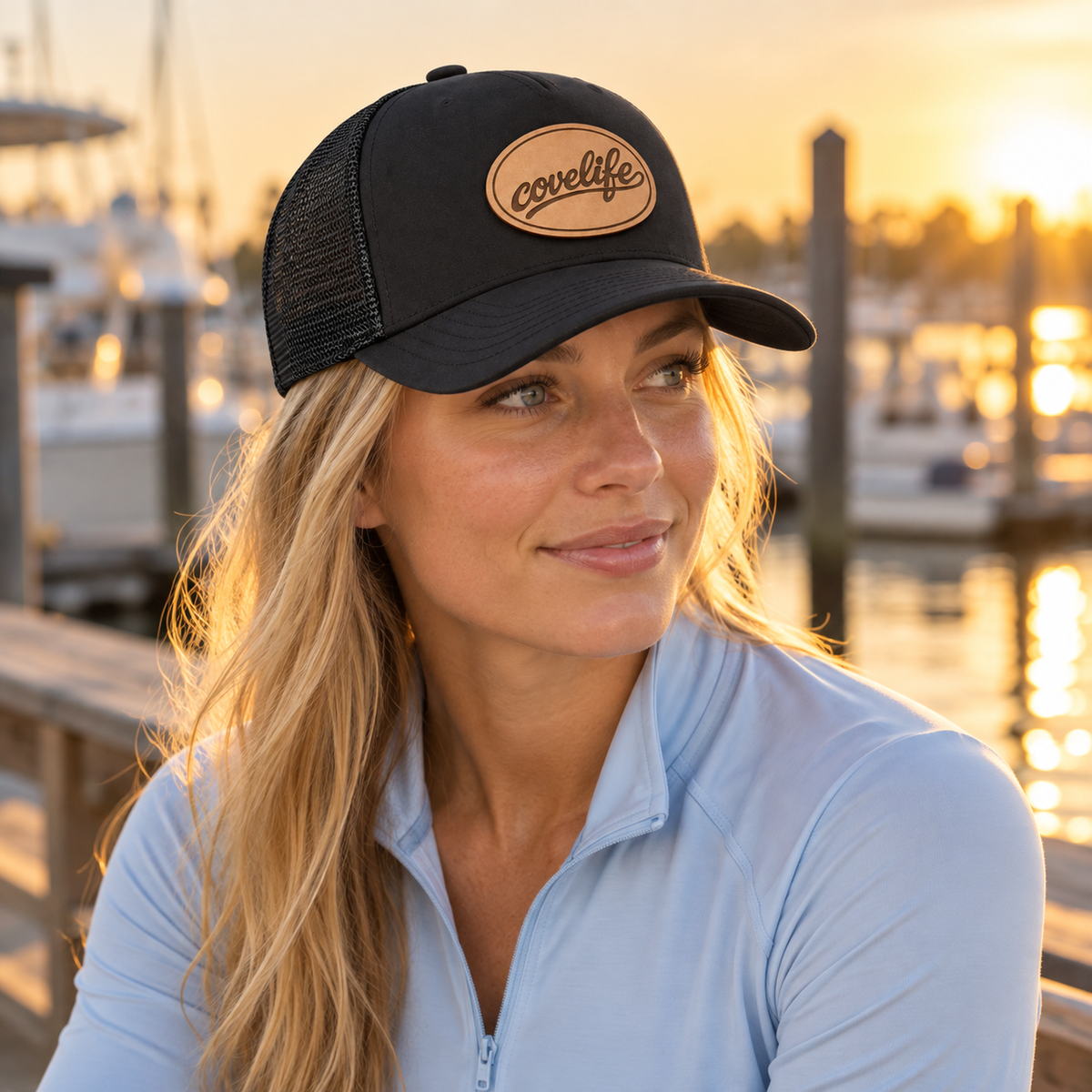 A woman wearing a black classic mesh hat with a tan oval patch that reads 'covelife' sits at a wooden dock with a marina and sunset in the background.