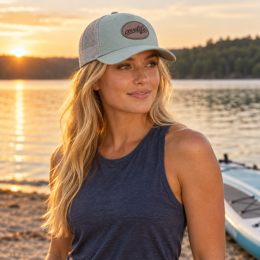A woman wearing a sage green classic mesh hat with a brown oval covelife logo leather patch, standing on a beach at sunset with water and a paddleboard in the background.