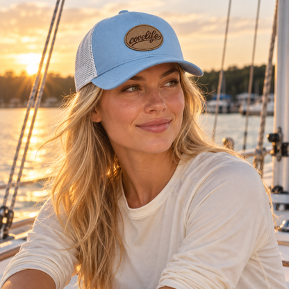 A woman on a boat at sunset wearing a sky blue classic mesh hat with a brown oval patch that reads covelife in script.