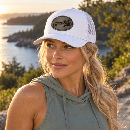 A woman outdoors wearing a white classic mesh covelife cap with a brown leather patch that reads 'covelife' and a sage green sleeveless hoodie, with a scenic lakeside background at sunset.