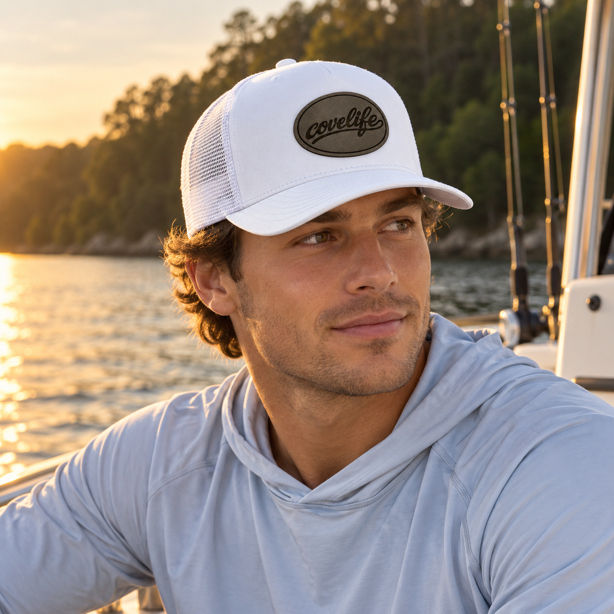A man wearing a white classic meshj hat with a brown oval 'covelife' patch and a light blue long-sleeve hoodie, photographed on a boat with water and trees in the background at sunset.