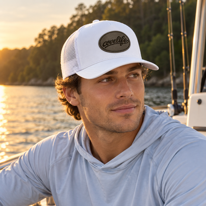 A man wearing a white classic meshj hat with a brown oval 'covelife' patch and a light blue long-sleeve hoodie, photographed on a boat with water and trees in the background at sunset.