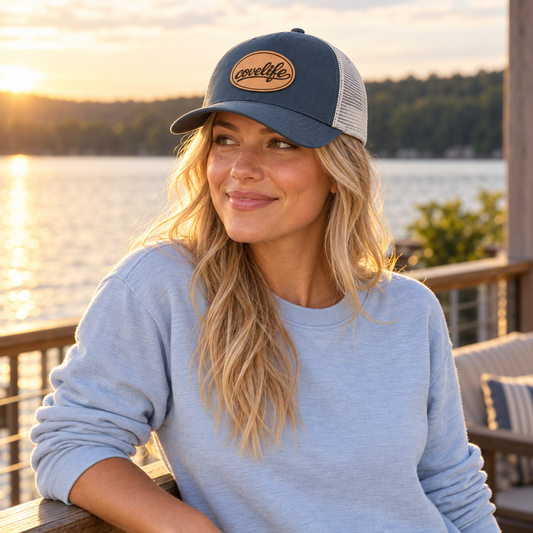 A woman wearing the Navy Classic Mesh hat with a heritage leather patch with covelife script.  sitting on a wooden deck with a lake and sunset in the background.