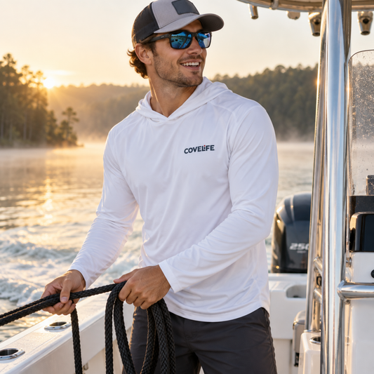 A man wearing a white long-sleeve CoveLife hoodie and sunglasses stands on a boat during sunset, holding a rope.