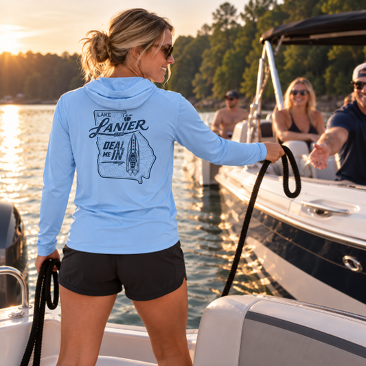 A woman on a boat at sunset wearing a light blue long-sleeve hooded shirt with a dark graphic print on the back that reads 'LAKE LANIER' and 'DEAL ME IN'.