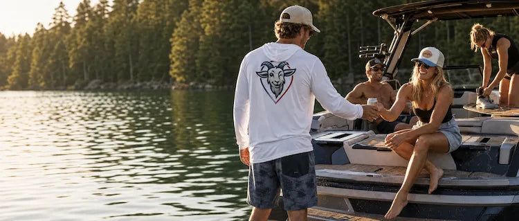 A man wearing a white long-sleeve solar tech UPF 50+ hoodie with brand goat mascot graphic on the back stands on a boat, shaking hands with a woman sitting on the boat. Other people are visible on the boat, and a forested lake is in the background during sunset.