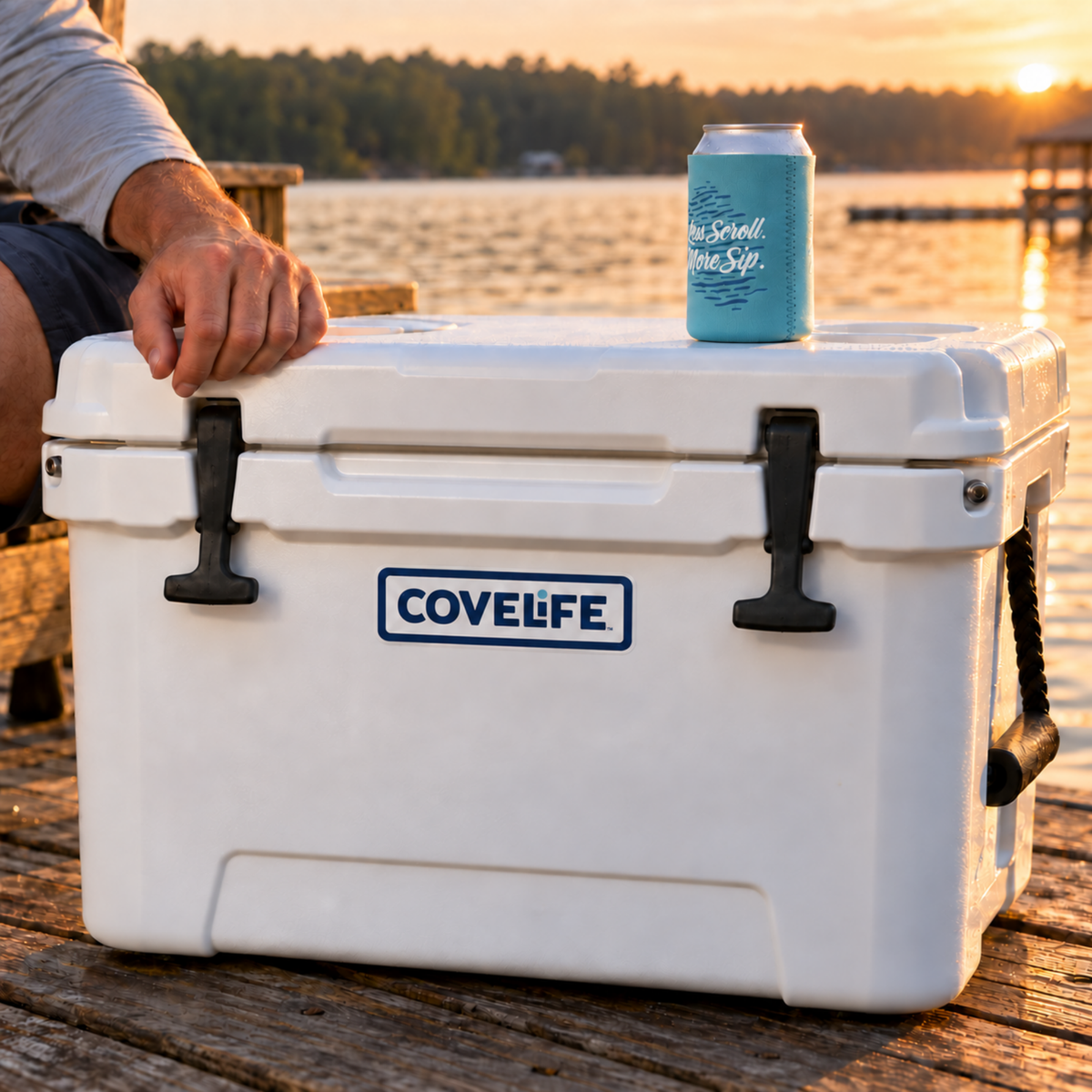 A white COVELIFE cooler sits on a wooden dock by the water at sunset, with a person's hand resting on the lid and a light blue can in a turquoise sleeve on top. The background shows a calm lake and trees in warm golden hour light.