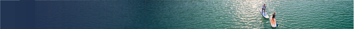 A wide, panoramic outdoor photograph of a calm lake or river with a small canoe or paddleboard visible near the upper-right area, under soft natural light.