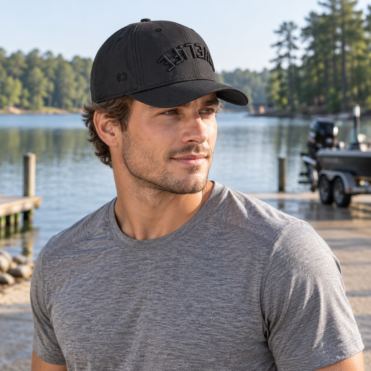 A man wearing a black baseball cap with the brand name 'COVELiFE' in black embroidery and a heather gray t-shirt, standing outdoors near a lake with a boat in the background.