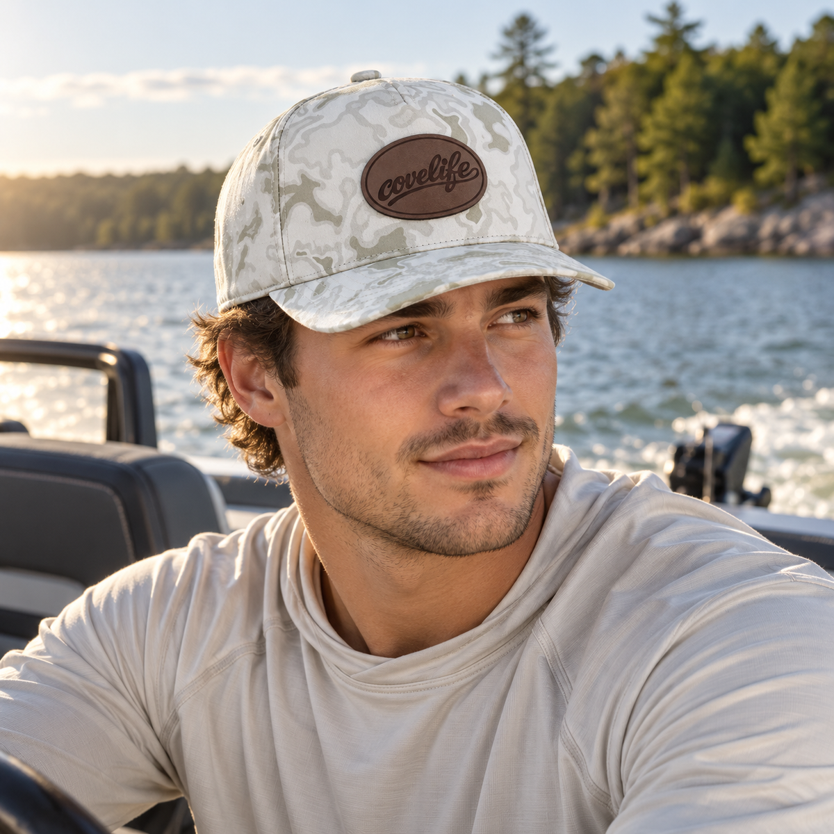A man wearing a light beige camouflage baseball cap with a brown oval covelife logo patch, sitting on a boat with water and trees in the background.