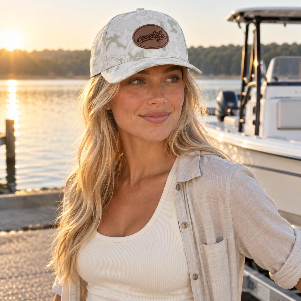 A woman wearing a white camouflage baseball cap with a brown oval patch that reads covelife stands by a lake at sunset, with a boat in the background.