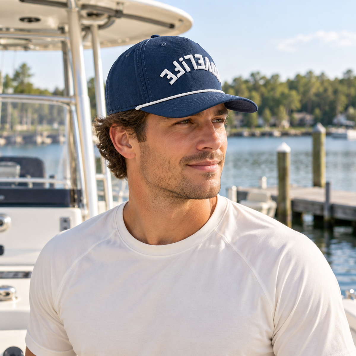 A man wearing a navy blue baseball cap with white COVELiFE front mark wearing a white t-shirt, photographed outdoors at a marina with water, boats, and trees in the background.