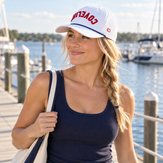 A woman standing on a dock by the water wearing a white baseball cap with red 'COVELiFE' lettering and a navy ribbed tank top, carrying a white tote bag.